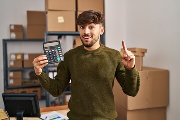 Wall Mural - Arab man with beard working at small business ecommerce holding calculator surprised with an idea or question pointing finger with happy face, number one