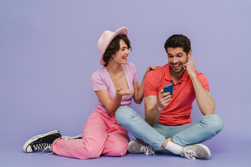 Wall Mural - Cheerful man and woman using smartphone while sitting isolated over purple background