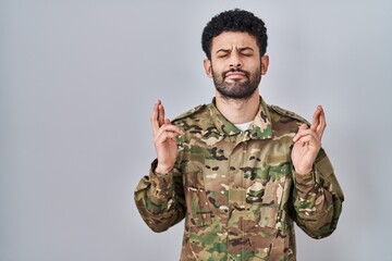 Canvas Print - Arab man wearing camouflage army uniform gesturing finger crossed smiling with hope and eyes closed. luck and superstitious concept.