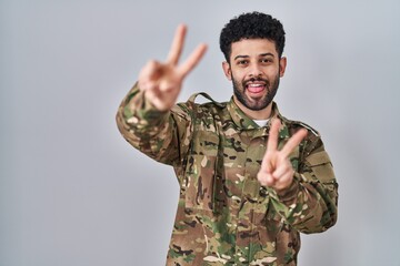 Canvas Print - Arab man wearing camouflage army uniform smiling with tongue out showing fingers of both hands doing victory sign. number two.