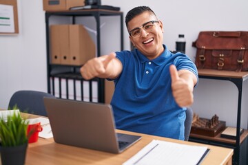 Wall Mural - Young hispanic man working at the office with laptop approving doing positive gesture with hand, thumbs up smiling and happy for success. winner gesture.