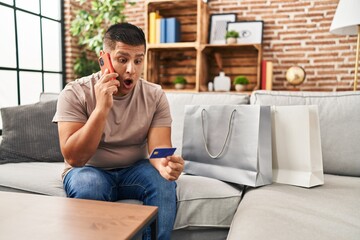 Canvas Print - Hispanic young man doing payment with credit card on the phone afraid and shocked with surprise and amazed expression, fear and excited face.