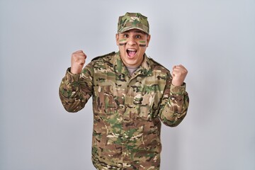 Wall Mural - Hispanic young man wearing camouflage army uniform celebrating surprised and amazed for success with arms raised and open eyes. winner concept.