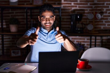 Sticker - Hispanic man with beard using laptop at night approving doing positive gesture with hand, thumbs up smiling and happy for success. winner gesture.