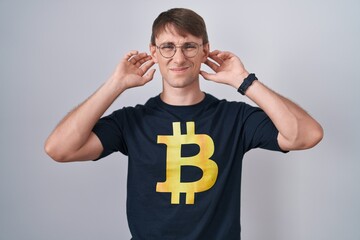 Poster - Caucasian blond man wearing bitcoin t shirt covering ears with fingers with annoyed expression for the noise of loud music. deaf concept.