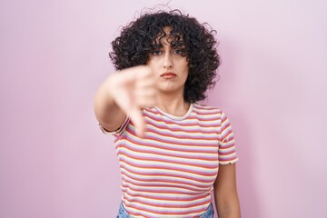 Wall Mural - Young middle east woman standing over pink background looking unhappy and angry showing rejection and negative with thumbs down gesture. bad expression.