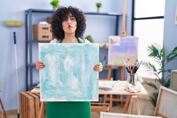 Sticker - Young brunette woman with curly hair holding painter canvas making fish face with mouth and squinting eyes, crazy and comical.