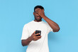 © Богдан Маліцький - puzzled african american guy in white t-shirt uses smartphone and makes facepalm on blue isolated background