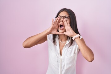 Wall Mural - Brunette young woman standing over pink background wearing glasses shouting angry out loud with hands over mouth