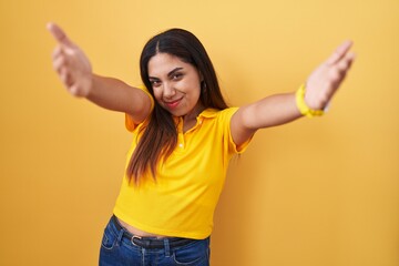 Wall Mural - Young arab woman standing over yellow background looking at the camera smiling with open arms for hug. cheerful expression embracing happiness.