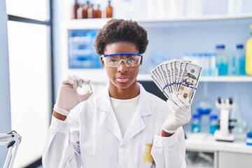 Wall Mural - African american woman working at scientist laboratory holding dollars relaxed with serious expression on face. simple and natural looking at the camera.