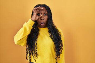 Sticker - African woman standing over yellow background doing ok gesture shocked with surprised face, eye looking through fingers. unbelieving expression.