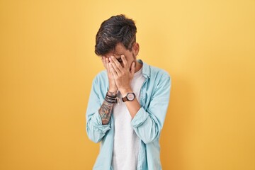 Wall Mural - Young hispanic man with tattoos standing over yellow background with sad expression covering face with hands while crying. depression concept.