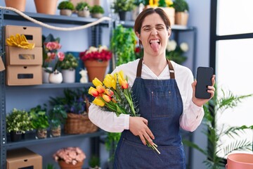 Sticker - Brunette woman working at florist shop holding smartphone sticking tongue out happy with funny expression.