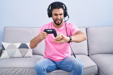 Wall Mural - Hispanic young man playing video game holding controller sitting on the sofa looking at the watch time worried, afraid of getting late