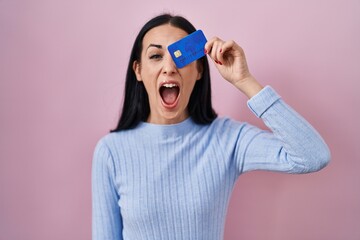 Sticker - Hispanic woman holding credit card over eye angry and mad screaming frustrated and furious, shouting with anger. rage and aggressive concept.