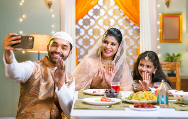Wall Mural - Happy smiling indian muslim family on video call on mobile phone during ramadan festival dinner or feast at home - conecept of distant relationship, festival celebration and cyberspace.