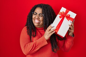 Sticker - Plus size hispanic woman holding gift winking looking at the camera with sexy expression, cheerful and happy face.