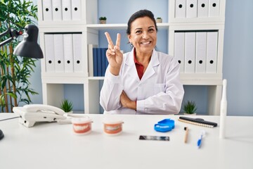 Poster - Hispanic mature dentist woman working at the clinic smiling with happy face winking at the camera doing victory sign. number two.