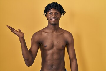 Canvas Print - Young african man with dreadlocks standing shirtless smiling cheerful presenting and pointing with palm of hand looking at the camera.