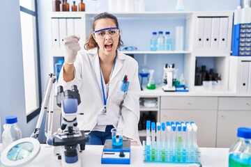 Wall Mural - Young hispanic woman working at scientist laboratory angry and mad raising fist frustrated and furious while shouting with anger. rage and aggressive concept.