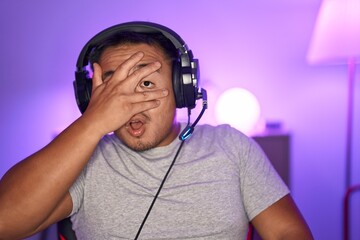 Poster - Chinese young man playing video games wearing headphones peeking in shock covering face and eyes with hand, looking through fingers afraid