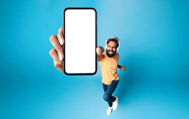 Wall Mural - Excited indian man in headphones demontrating smartphone with blank screen while jumping up, mockup, above view