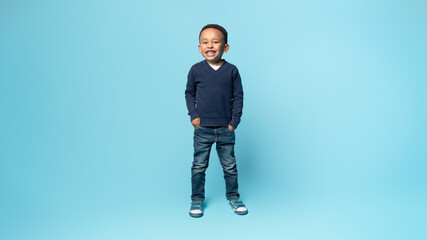 Wall Mural - Full length shot of african american little boy posing holding hands in pockets, standing on blue background, panorama