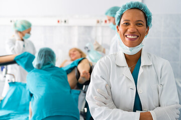 Portrait of successful smiling African American female doctor gynecologist with surgical cap and uniform looking at camera standing in front of medical team and woman in labor.