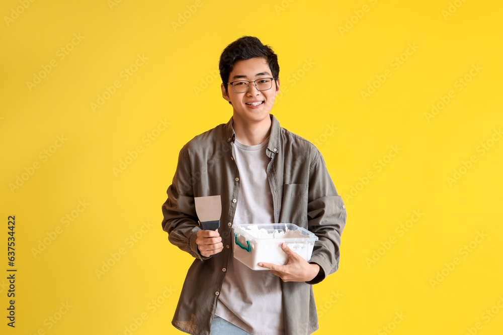 Young Asian man with putty knife and plaster on yellow background