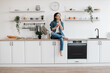 © sofiko14 - Thoughtful woman with mobile in hands sitting on kitchen