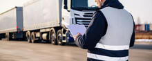 A Male Worker Wearing A White Ondown Shirt With The CrossBorder Logo On The Chest Beige Trousers And Black Boots. He Is Checking The Documentation Of A Truck Driver Passing Through The CrossBorder