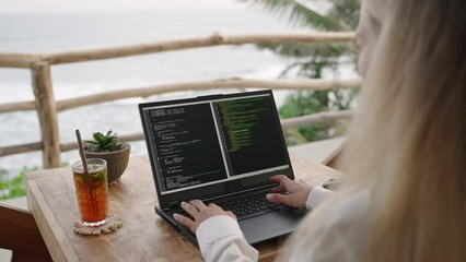 Female soft dev with laptop on remote work day in tropical cafe. Software developer writes program code on computer screen in coworking restaurant with sea view on sunset. Woman coding by ocean.