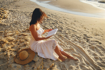 Romantic girl in a white dress reads a book sitting on a stone on the beach. View from above.