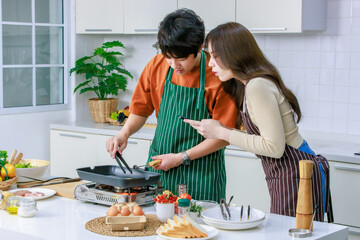 Asian young lover couple husband and wife in casual outfit with apron standing smiling while man cooking frying food with pan woman taking photo with smartphone in full decorated modern kitchen