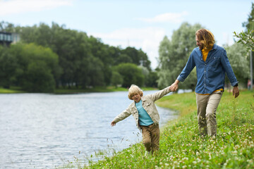 Full length portrait of father and son walking together by lake in green forest, copy space