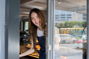 Young adult Asian female barista welcoming a customer walking into the coffee shop with a friendly smile and warm welcome. Small Business owner in Asia. Thai language sign on the door is to slide