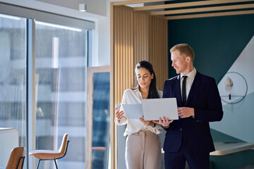 Two busy diverse coworkers discussing project walking in office with laptop and documents. International business team people colleagues standing having teamwork conversation. Authentic shot