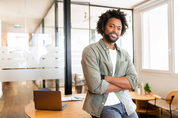 Successful african-american curly male entrepreneur stands and looks at the camera with arms folded, positive smiling freelancer man in casual shirt in coworking space, ceo, manager, start-up owner