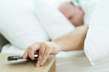 Lazy oversize fat man trying to turn off or snooze the alarm clock on smartphone while he sleeping on the bed in morning, a lazy lifestyle concept. Man looking the time on alarm clock on smartphone.