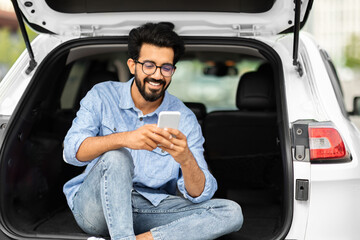 Wall Mural - Happy indian guy sitting in open car trunk, using smartphone