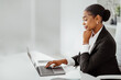Confident black businesswoman looking at laptop and smiling, sitting at workplace in office, side view, free space