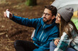 Cheerful young caucasian couple in jackets enjoy travel vacation with tent, make selfie on smartphone