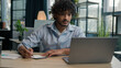 © Yuliia - Arabian Indian latin man businessman guy write business notes ideas in notebook managing corporate tasks writing schedule with pen on desk male worker employee working with laptop computer in office