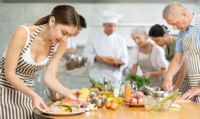 Interested engrossed young woman honing cooking skills during group culinary classes, garnishing and seasoning finished dish with sauce before presentation