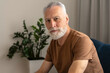 © Maria Vitkovska - Handsome pensioner wearing brown t shirt, sitting on sofa in living room, flowerpot on background
