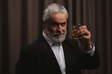 Wall Mural - Senior man in formal suit holding glass of whiskey with ice cubes on brown background