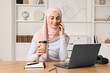 © New Africa - Muslim woman with cup of coffee talking on smartphone near laptop at wooden table in room