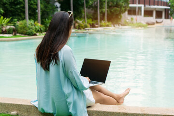 Freelancer using laptop working remotely near swimming pool. Young Asian traveler woman working on computer during her summer holiday. Work from anywhere, Technology and lifestyle concept, copy space