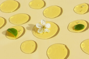 Wall Mural - Drops of transparent cosmetic gel or serum yellow with a white flower and green leaves on a beige background. The concept of natural cosmetics. Top view. Selective focus.